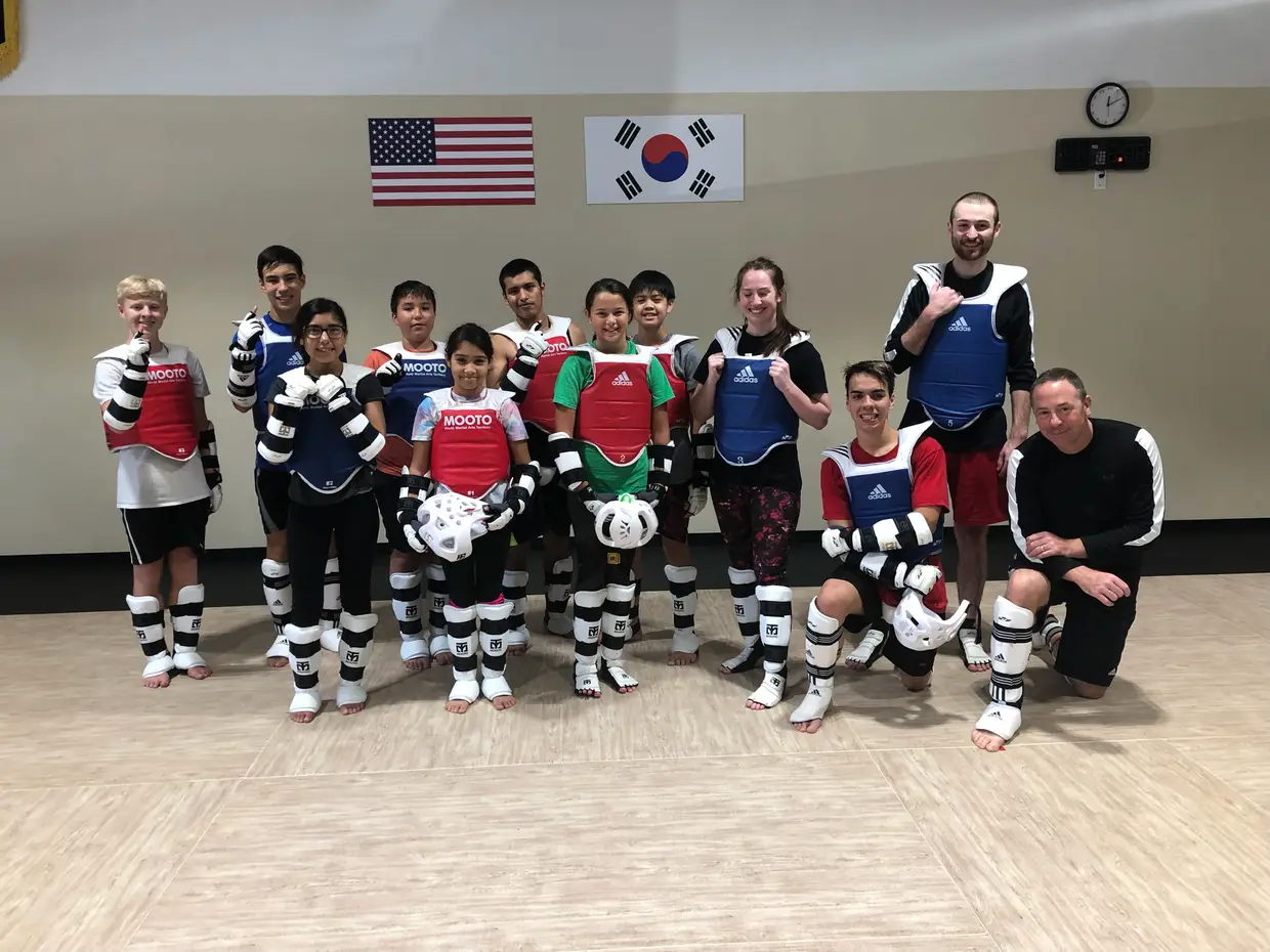 Force of One students in full sparring gear — red and blue hogu, shin and forearm guards — gathered in the dojang beneath the American and Korean flags
