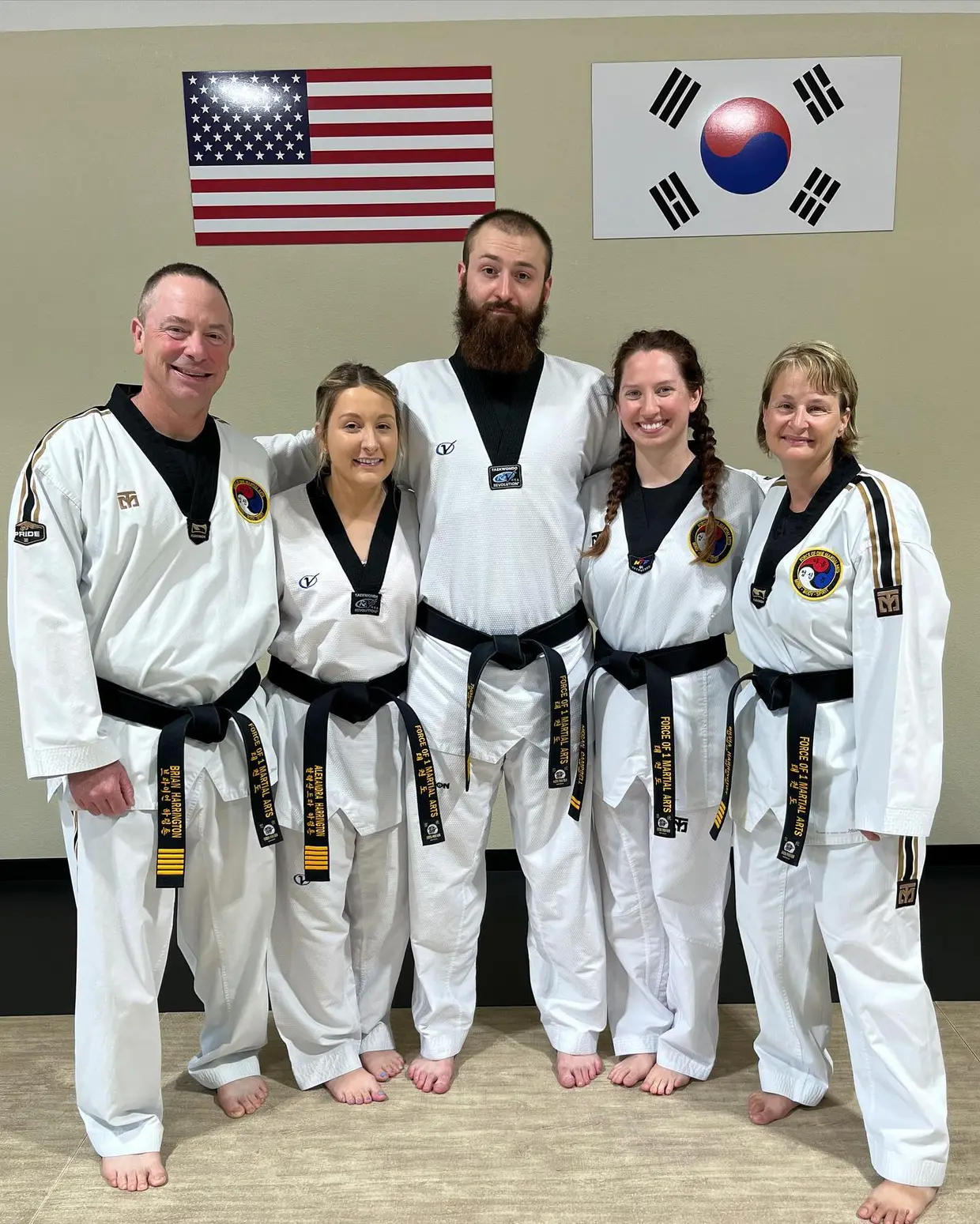 The Force of One instructors in white dobok and black belts, standing together in the dojang beneath the American and Korean flags