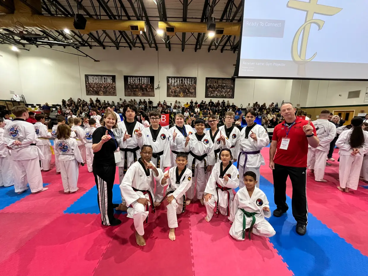 The Force of One team and their coaches on the red and blue competition mats at a Taekwondo tournament, surrounded by competitors from other schools